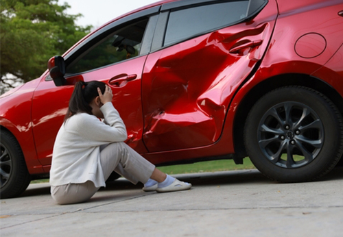 A woman sits on the ground next to her red car with a severely dented rear door, clutching her head in distress after a car accident.